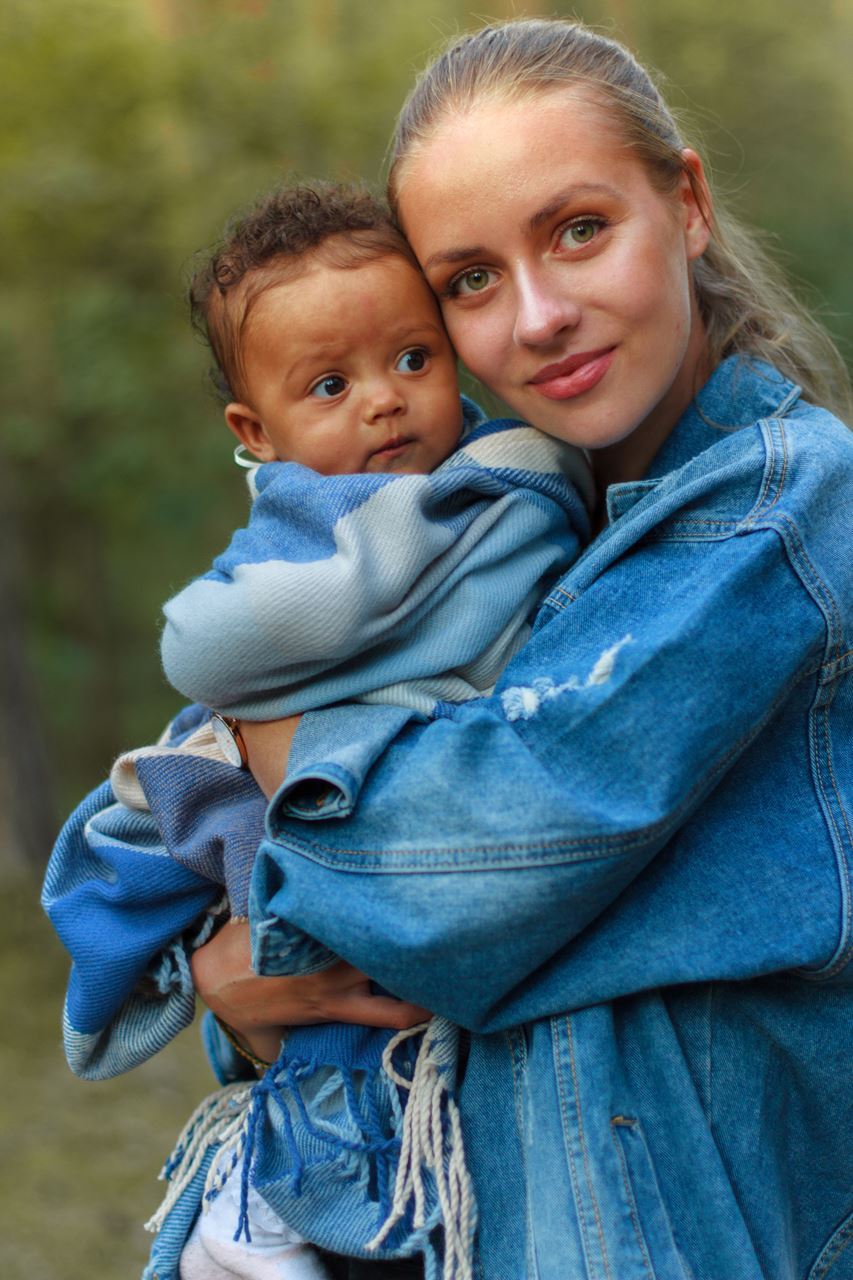 photo of woman in denim jacket holding an infant wrapped in a plaid blanket. Both are looking at the camera. Photo by Humphrey Muleba on Unsplash.